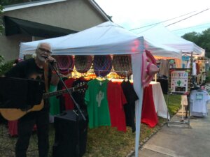 Older man signing with guitar with a white tent and vendor items